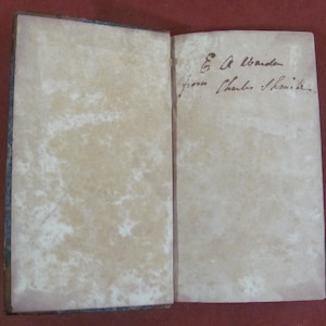 May include: An open antique book with aged, off-white paper pages showing significant foxing and discolouration. The right page features handwritten cursive ink text that reads E A Warden from Charles Smith.
