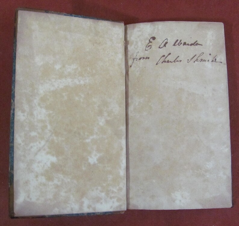 May include: An open antique book with aged, off-white paper pages showing significant foxing and discolouration. The right page features handwritten cursive ink text that reads E A Warden from Charles Smith.