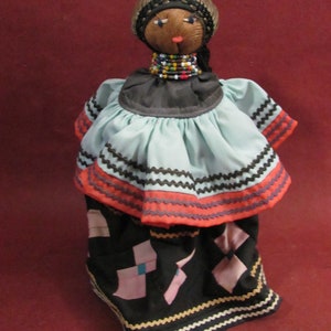 May include: Handmade doll with a brown head, wearing a black head covering and a beaded necklace. The doll has a light blue top with orange and black trim, and a black skirt with pink and blue geometric shapes. The doll is standing against a red background.