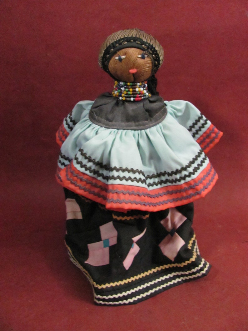 May include: Handmade doll with a brown head, wearing a black head covering and a beaded necklace. The doll has a light blue top with orange and black trim, and a black skirt with pink and blue geometric shapes. The doll is standing against a red background.