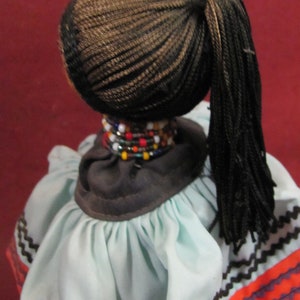 May include: A doll with dark braided hair pulled back in a ponytail. The doll wears a light blue dress with a dark brown collar and colorful beaded necklace. The dress has a zigzag pattern in brown, orange, and black.