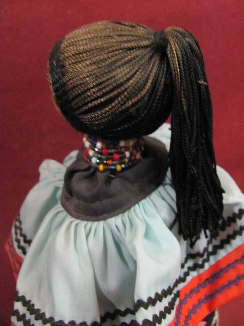 May include: A doll with dark braided hair pulled back in a ponytail. The doll wears a light blue dress with a dark brown collar and colorful beaded necklace. The dress has a zigzag pattern in brown, orange, and black.