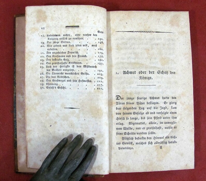 Op de afbeelding: Een open antiek boek met vergeelde, gebroken witte papieren pagina's met zwarte gedrukte Duitse tekst. De linkerpagina toont een genummerde inhoudsopgave, terwijl de rechterpagina een hoofdstuktitel en een tekstgedeelte weergeeft.