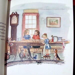 May include: Three girls are gathered around a table in a kitchen, making small pies. They are wearing dresses and aprons. There are two cats and a dog in the room. The room has a clock on the wall, a window with a red curtain, and a picture on the wall.