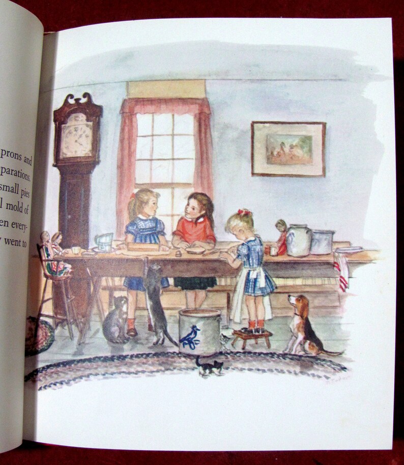 May include: Three girls are gathered around a table in a kitchen, making small pies. They are wearing dresses and aprons. There are two cats and a dog in the room. The room has a clock on the wall, a window with a red curtain, and a picture on the wall.