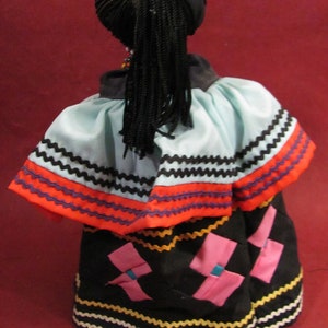 May include: A doll with black braided hair and a black head covering. The doll is wearing a light blue and orange top with black and white trim. The doll's skirt is black with pink and blue accents and yellow and white trim.