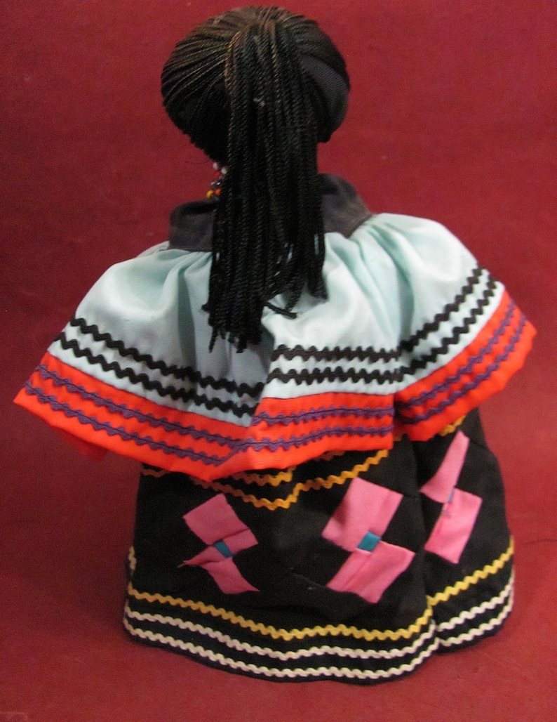 May include: A doll with black braided hair and a black head covering. The doll is wearing a light blue and orange top with black and white trim. The doll's skirt is black with pink and blue accents and yellow and white trim.