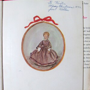 May include: A vintage illustration of a young girl wearing a brown dress and a white lace collar. The illustration is framed in a gold oval and surrounded by a red ribbon. The text "To Kustin Merry Christmas, 1970. From Mother" is written in blue ink on the page.