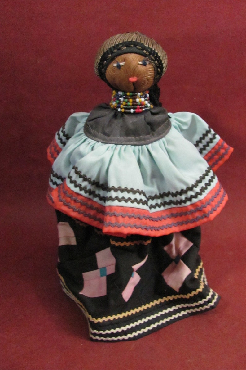 May include: A handmade doll with a dark complexion, wearing a light blue dress with orange and black trim. The doll has a beaded necklace and a woven head covering. The skirt is black with pink and blue geometric shapes.