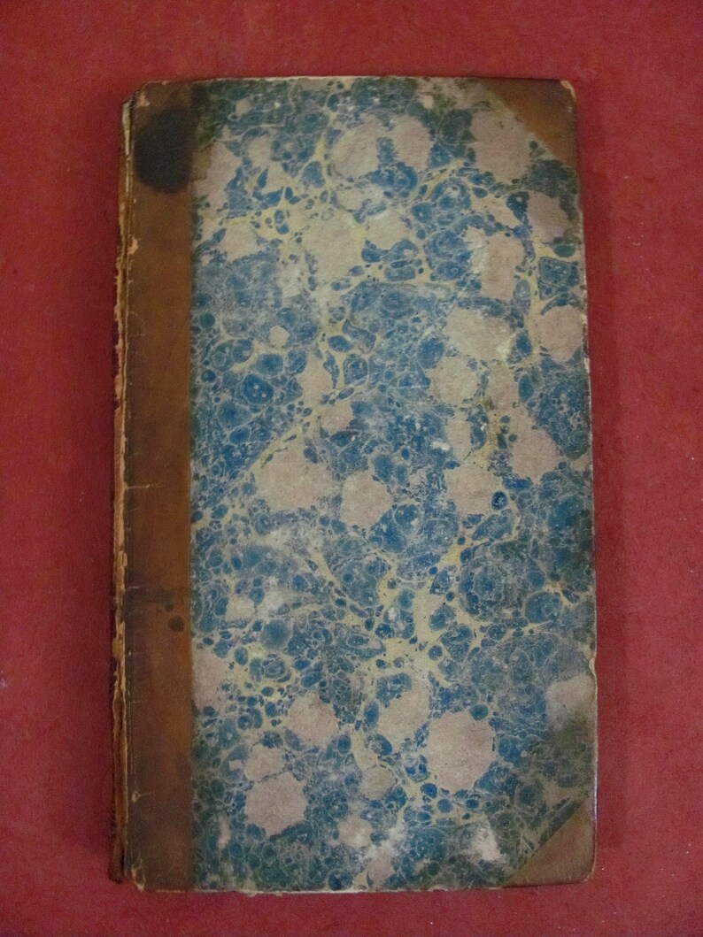 Op de afbeelding: Een rechthoekig antiek boek met een gemarmerde kaft met een patroon van blauwe en beige wervelingen. De rug en de linkerkant van de kaft zijn gebonden in versleten bruin leer, met tekenen van ouderdom en slijtage langs de randen.