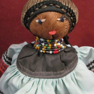 May include: A handmade doll with a brown face, stitched features, and a woven brown cap with a black trim. It wears a black collar adorned with colorful beads and a light blue dress with black and orange trim.