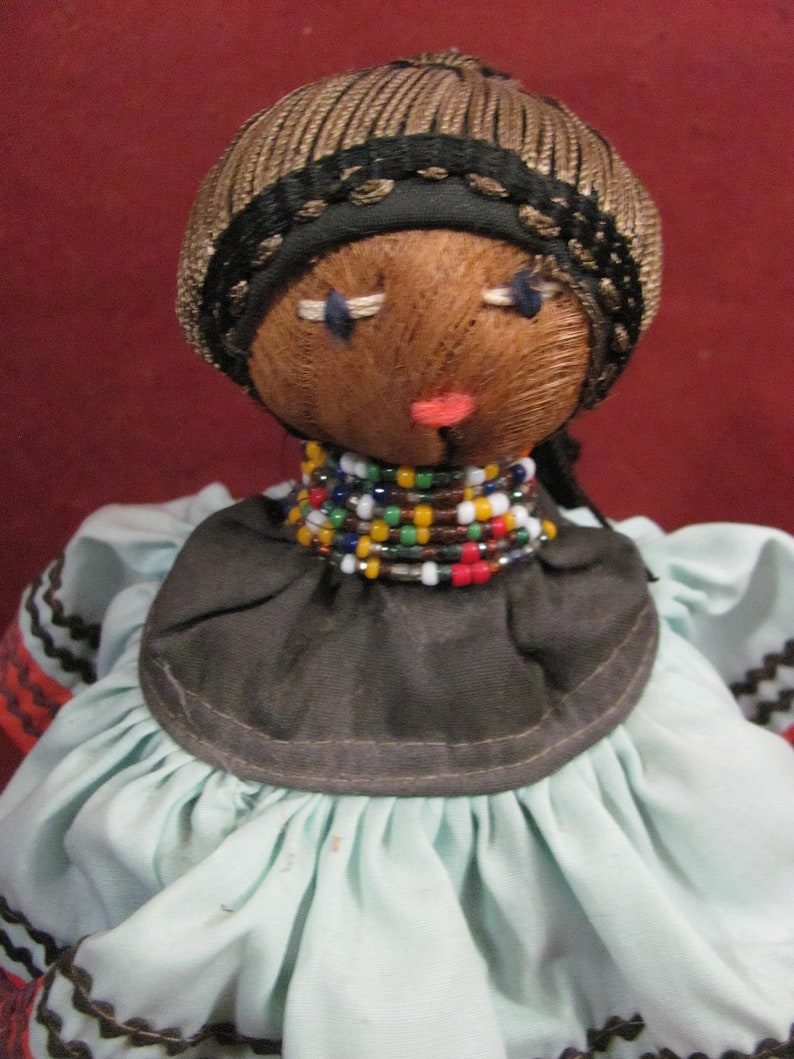 May include: A handmade doll with a brown face, stitched features, and a woven brown cap with a black trim. It wears a black collar adorned with colorful beads and a light blue dress with black and orange trim.