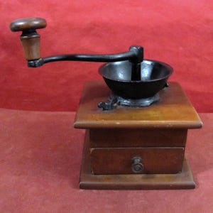 Può includere: Un'antica macinacaffè in legno con una ciotola e una manovella in metallo nero. Il macinacaffè ha un piccolo cassetto e un manico in legno sulla manovella. Il legno è marrone caldo, lo sfondo è rosso.