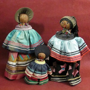 May include: Three handmade dolls with coconut shell heads and beaded necklaces. The dolls wear colorful fabric dresses with intricate trim. The largest doll has a wide-brimmed hat. The dolls are arranged on a maroon background.
