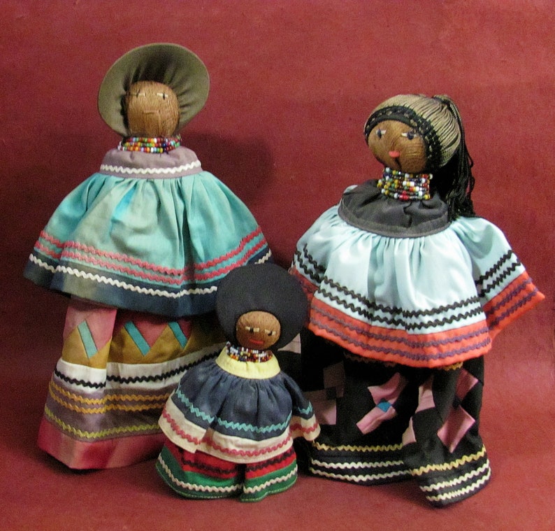 May include: Three handmade dolls with coconut shell heads and beaded necklaces. The dolls wear colorful fabric dresses with intricate trim. The largest doll has a wide-brimmed hat. The dolls are arranged on a maroon background.