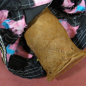 May include: A close-up shot of a brown, woven basket-like object with a flared shape. The basket is set against a backdrop of black fabric adorned with pink and blue fabric accents. White stitching outlines the fabric.