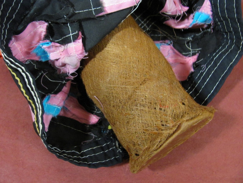 May include: A close-up shot of a brown, woven basket-like object with a flared shape. The basket is set against a backdrop of black fabric adorned with pink and blue fabric accents. White stitching outlines the fabric.