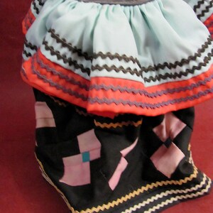 May include: A doll's traditional dress with a light blue top and a black skirt. The top has a ruffled design with orange and brown trim. The skirt features pink and teal geometric shapes and is trimmed with a yellow and white zigzag pattern.