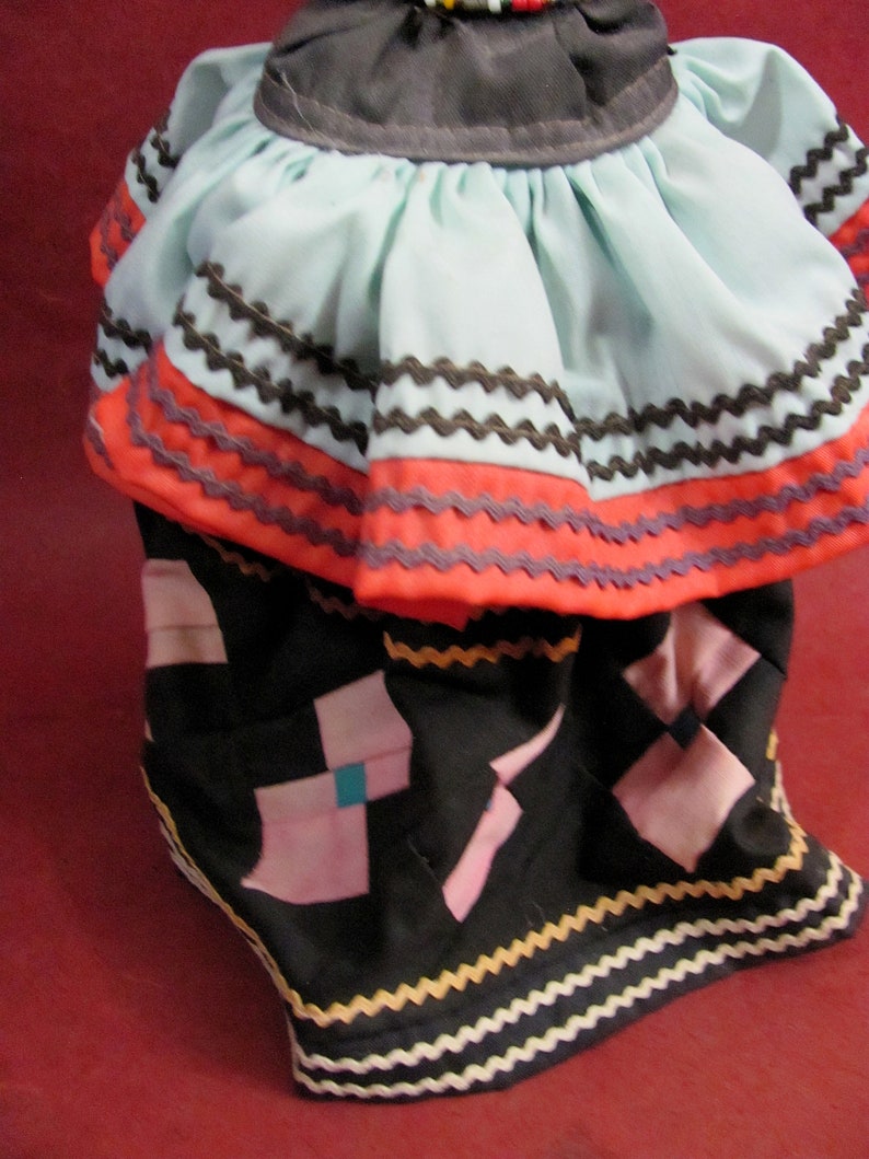 May include: A doll's traditional dress with a light blue top and a black skirt. The top has a ruffled design with orange and brown trim. The skirt features pink and teal geometric shapes and is trimmed with a yellow and white zigzag pattern.
