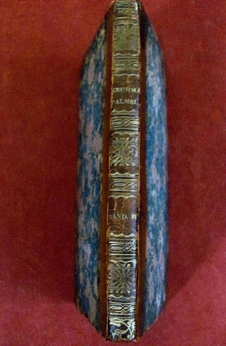 Op de afbeelding: Een antiek boek met een bruine lederen rug voorzien van goudkleurige decoratieve patronen en de tekst KRUMMA PALMEL BAND II. De boekomslagen hebben een blauw en zwart gemarmerd patroon met een versleten, getextureerd uiterlijk.