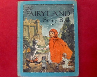 Op de afbeelding: Vintage "The Fairyland Story Book" met een blauwe kaft. De illustratie toont een meisje in een rode cape, een kasteel en verschillende dieren. De achtergrond toont een bergketen, wat een sprookjesscène creëert.