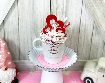 May include: White ceramic mug with a red and white whipped cream topping, red paper hearts, and a red and white striped straw. The mug has the word "love" written on it. The mug is sitting on a white cake stand with a pink doily.