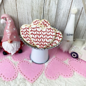 May include: A white cake stand with a black rim holds a stack of heart-shaped cookies decorated with a red and white knit pattern. The cookies are on a white background with pink heart-shaped doilies.