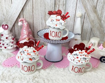 May include: Three white mugs with red "XOXO" text, topped with faux whipped cream, red heart decorations, and straws. The mugs are on a pink surface with Valentine's Day themed decorations in the background.