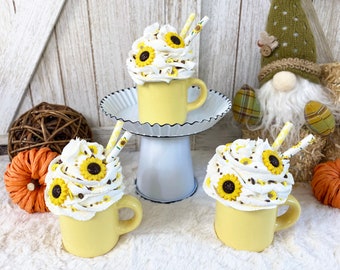 May include: Three yellow mugs with white whipped cream topped with chocolate sprinkles and miniature sunflowers. The mugs are on a white pedestal and a white fuzzy surface.
