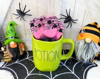 Könnte beinhalten: Eine grüne Tasse mit dem Wort "POTION" darauf, gefüllt mit rosa und schwarzen Halloween-Kekse. Die Tasse steht auf einer schwarzen Oberfläche mit Spinnennetz-Muster. Auf beiden Seiten der Tasse stehen zwei Gnome, einer mit einer Hexenmütze und der andere mit einer gestreiften Mütze.
