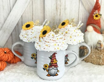 May include: Three white ceramic mugs with a fall theme. Each mug has a white whipped cream topping with a yellow sunflower and gold glitter. The mug in the center has a gnome illustration with a red hat and a sunflower.