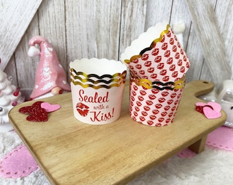 May include: Set of four cupcake liners with a scalloped edge and gold trim. The liners are white with a red lip print and one liner has the text "Sealed with a Kiss!" in red and gold.