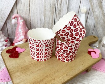 May include: Cupcake liners with scalloped edges. One set features red "XOXO" text on a white background, while another displays a red lipstick kiss pattern. The liners are on a wooden cutting board.
