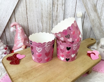 May include: Pink cupcake liners with a heart pattern. The liners are stacked on a wooden surface.