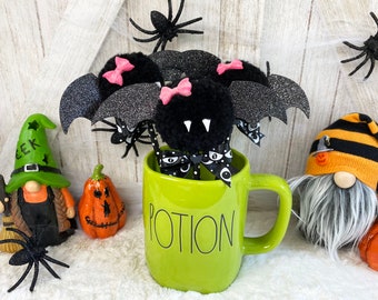 Könnte beinhalten: Eine grüne Tasse mit dem Wort "POTION" darauf. Die Tasse ist gefüllt mit schwarzen und pinken Fledermaus-förmigen Pompons mit Glitzer. Die Tasse ist umgeben von Halloween-Dekorationen, darunter Gnome, Kürbisse und Spinnen.