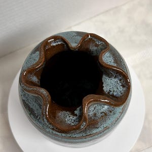 May include: A handmade ceramic vase with a unique ruffled rim. The vase features a two-tone glaze, with a blue-grey body and a rich brown rim. The vase sits on a white circular base, showcasing its handcrafted details.