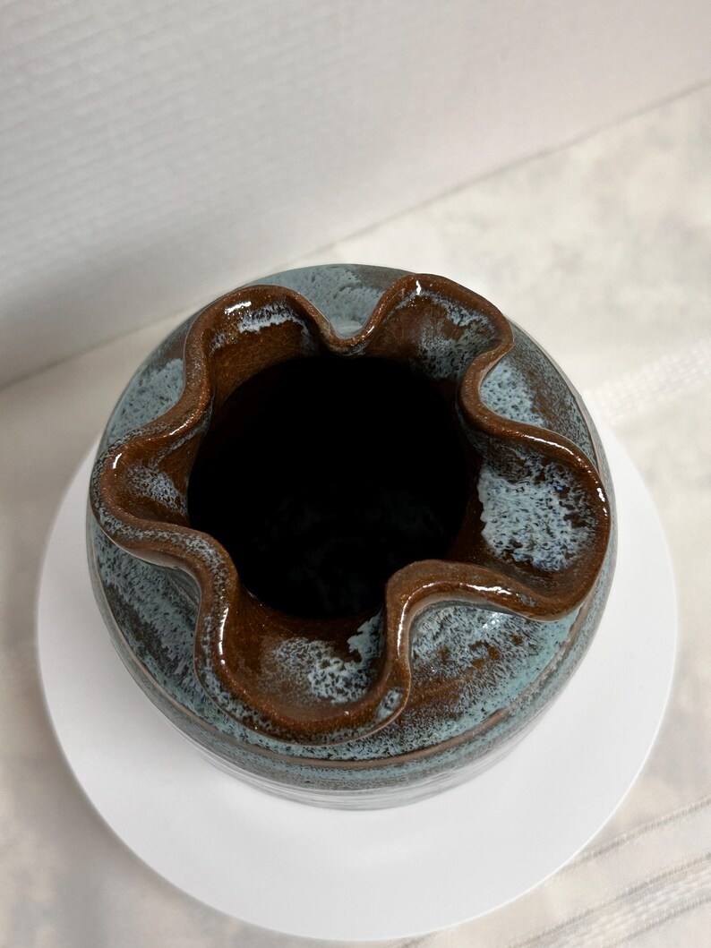 May include: A handmade ceramic vase with a unique ruffled rim. The vase features a two-tone glaze, with a blue-grey body and a rich brown rim. The vase sits on a white circular base, showcasing its handcrafted details.