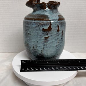 May include: A ceramic vase with a ruffled rim, featuring a blend of blue and brown hues. The vase has a cylindrical body and a unique, textured surface. A ruler is placed next to the vase for scale.