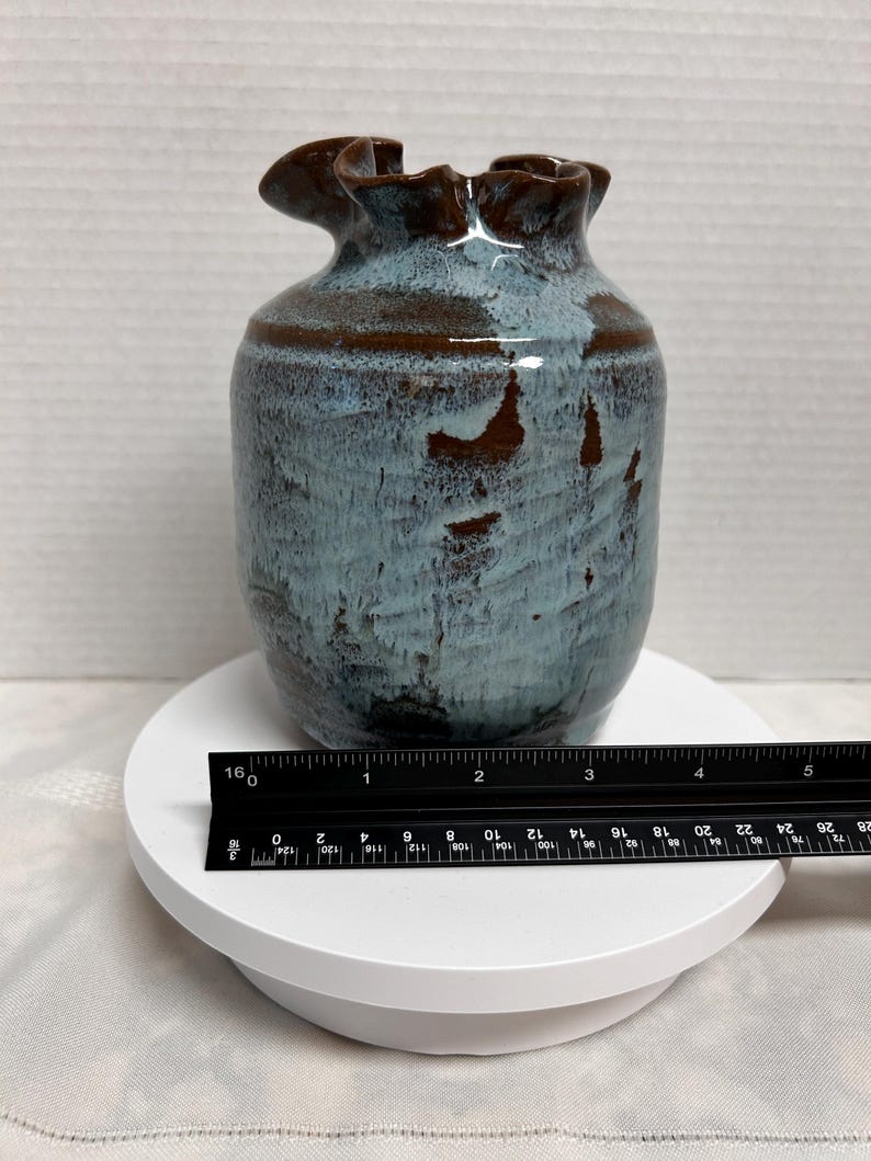May include: A ceramic vase with a ruffled rim, featuring a blend of blue and brown hues. The vase has a cylindrical body and a unique, textured surface. A ruler is placed next to the vase for scale.