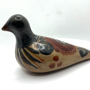 May include: A ceramic bird figurine with a black head and beak, and a body painted in shades of brown, tan, and black. The bird has decorative patterns on its wings and body. The figurine is resting on a white surface.