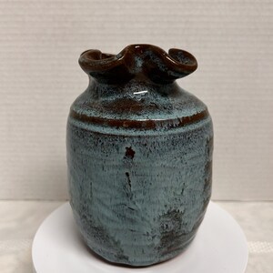 May include: A small, handcrafted ceramic vase with a unique shape and color. The vase features a dark brown, ruffled rim and a body with a mottled blue-green glaze. A brown band encircles the middle of the vase.