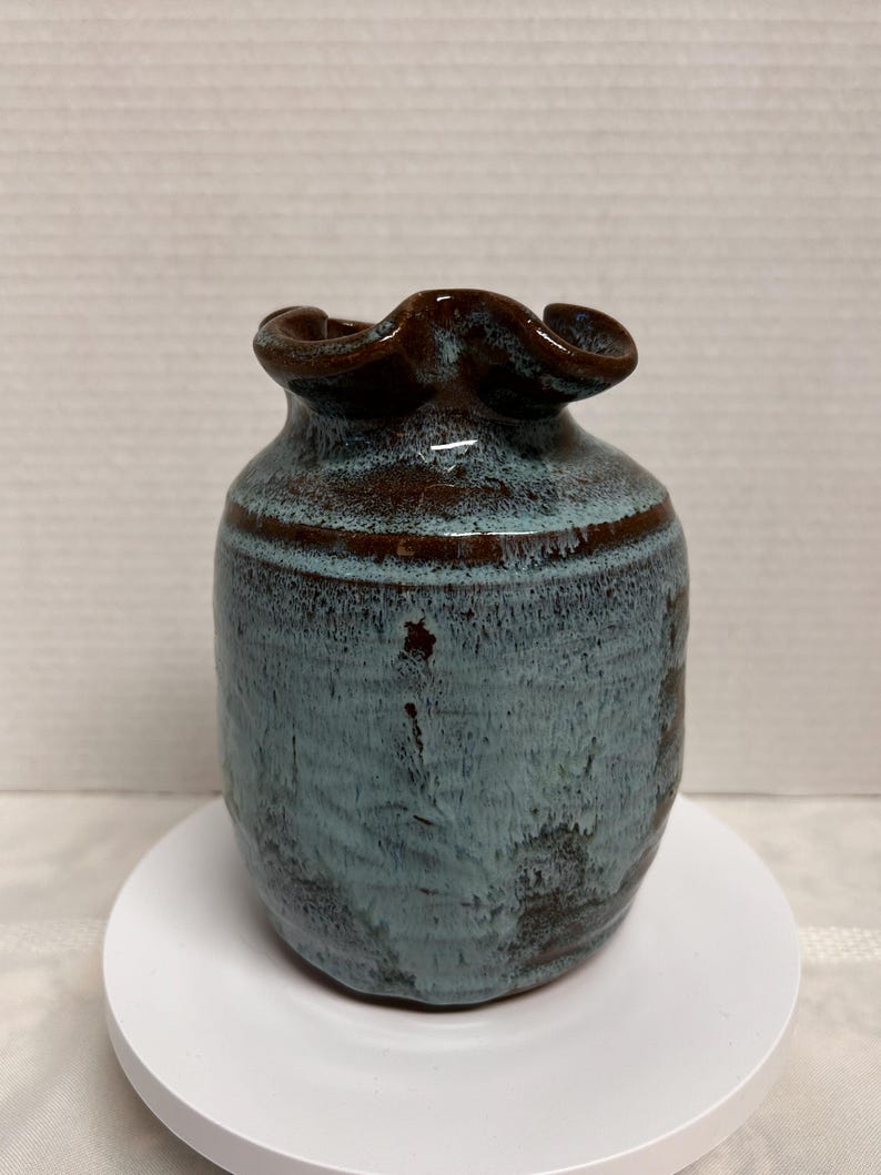 May include: A small, handcrafted ceramic vase with a unique shape and color. The vase features a dark brown, ruffled rim and a body with a mottled blue-green glaze. A brown band encircles the middle of the vase.
