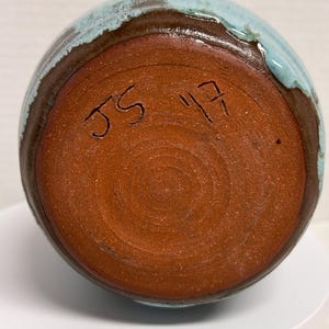 May include: A handmade ceramic vase with a round shape. The vase features a brown base and rim, with a turquoise blue glaze on the top. The bottom of the vase has the handwritten initials "JS" and the number "17".