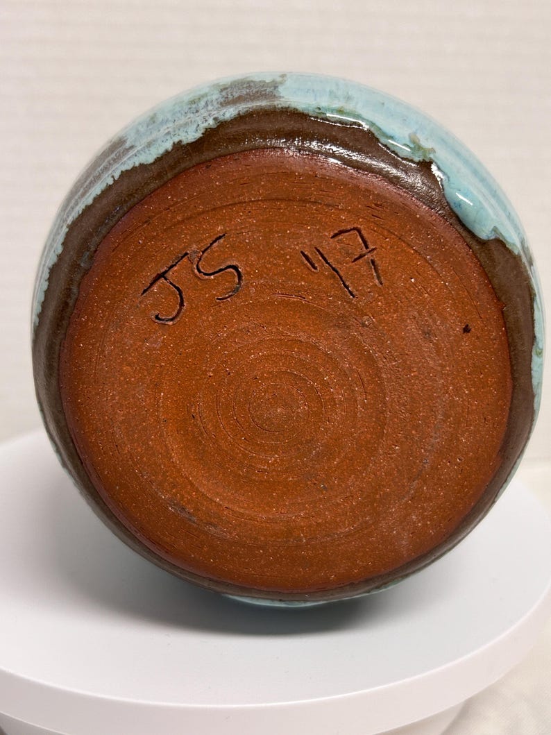 May include: A handmade ceramic vase with a round shape. The vase features a brown base and rim, with a turquoise blue glaze on the top. The bottom of the vase has the handwritten initials "JS" and the number "17".