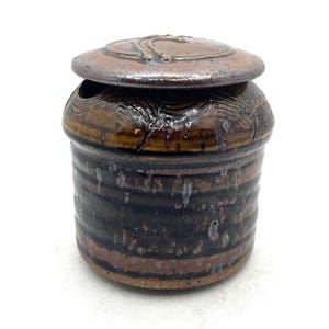May include: A cylindrical ceramic jar with a matching lid, featuring a dark brown and tan glaze with horizontal ribbed textures and subtle metallic-toned speckling. The lid has a slightly domed shape with carved linear details on the surface.