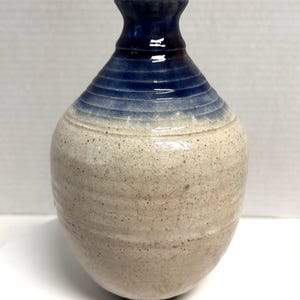 May include: A handmade ceramic vase with a speckled, off-white body and a dark blue neck. The vase features a gradient of blue hues around the upper portion, with horizontal ridges adding texture. The vase has a rounded shape.