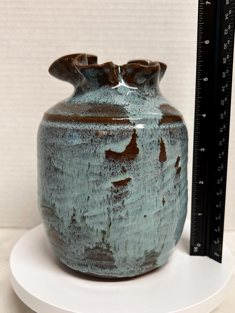 May include: A handcrafted ceramic vase with a unique ruffled rim. The vase features a blend of blue and brown glaze, creating a textured, organic appearance. The vase is approximately 6 inches tall.