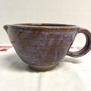 May include: A handmade ceramic mixing bowl with a handle and a spout. The bowl has a mottled brown and purple glaze. The handle is a rounded loop. The bowl sits on a small, unglazed base.