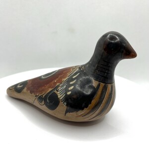 May include: A ceramic bird figurine with a brown beak and a black head. The body is tan with brown, black, and white painted details. The bird is resting on a white surface.