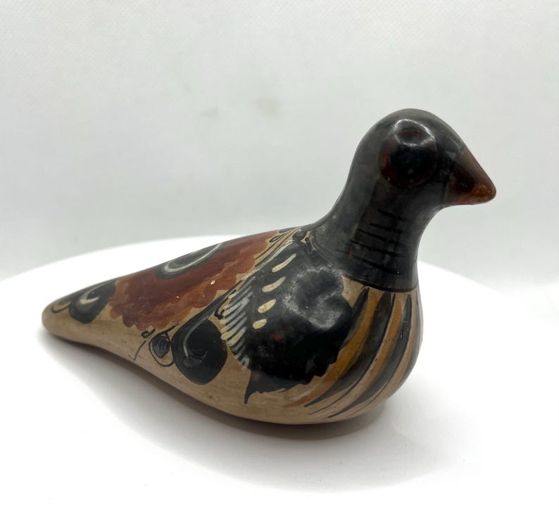 May include: A ceramic bird figurine with a brown beak and a black head. The body is tan with brown, black, and white painted details. The bird is resting on a white surface.